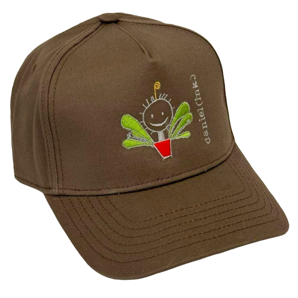 Walnut snapback cap featuring an embroidered image of a happy humanoid pot plant. The plant person is smiling and has a red plant pot-shaped torso with green leaves sprouting from the torso and a gold antenna sprouting from the top of its head. The name daniel(ink.) is embroidered in silver thread next to the plant person. 