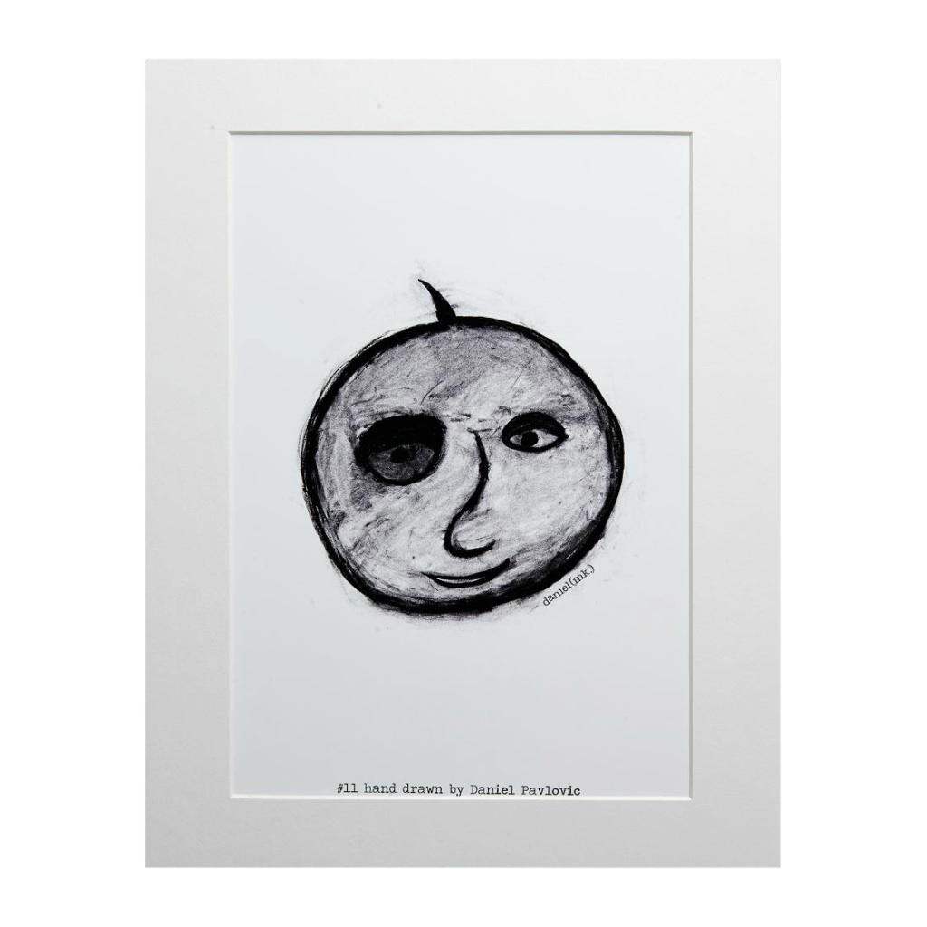 Original print of Design #11 Apple Face. Apple Face is a hand drawn charcoal sketch of an apple with a face in black and white. The design is printed on white backing board and displayed in a white frame. 